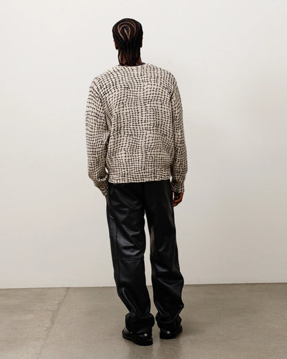 A model viewed from behind wearing a beige and black patterned knit sweater and black leather pants, standing against a plain off-white wall on a concrete floor.